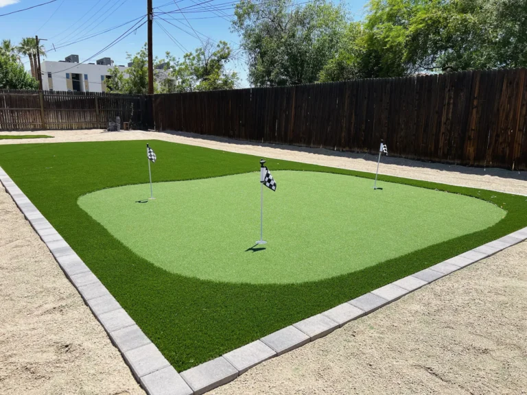 A backyard putting green with three small checkered flags on poles, surrounded by lush artificial turf installations and a border of gray bricks, sits next to a wooden fence and trees under the sunny Texas sky.