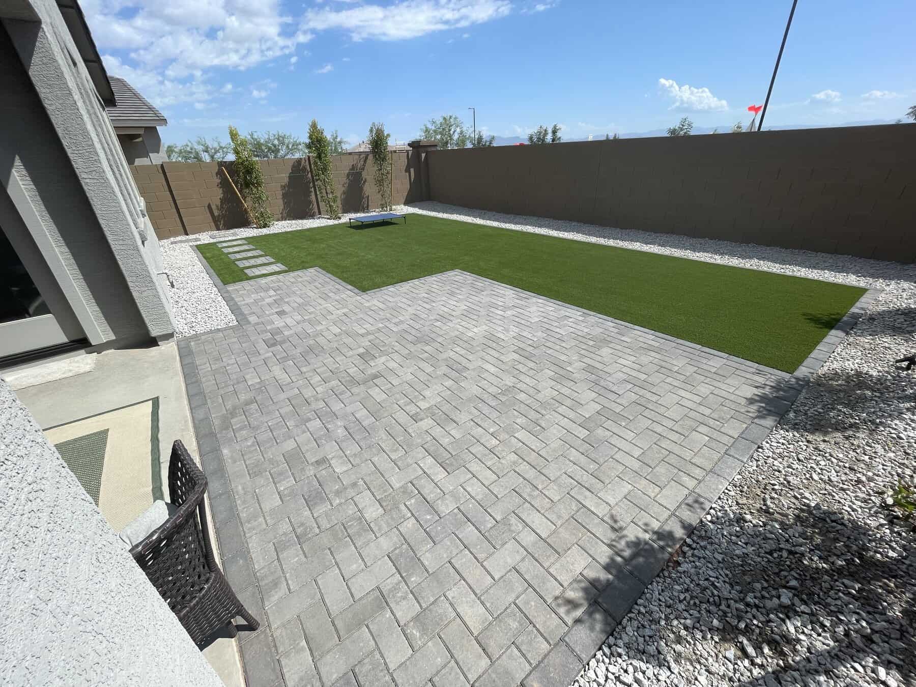 A backyard with a swimming pool, artificial grass, two beige lounge chairs, block wall fencing, and an air conditioning unit, adjacent to a house. The sky is cloudy and neighboring houses are visible.