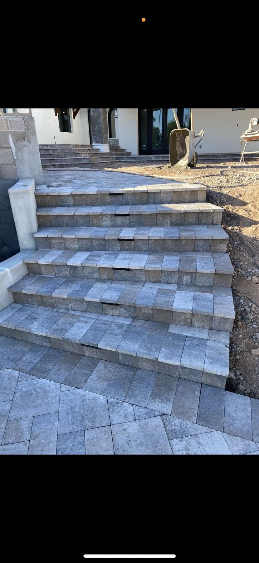 Outdoor stone steps under construction, featuring light gray rectangular pavers in a staggered pattern leading to a house. Dirt and a wheelbarrow are visible to the right—work expertly handled by a Pavers Installer Texas specialist.