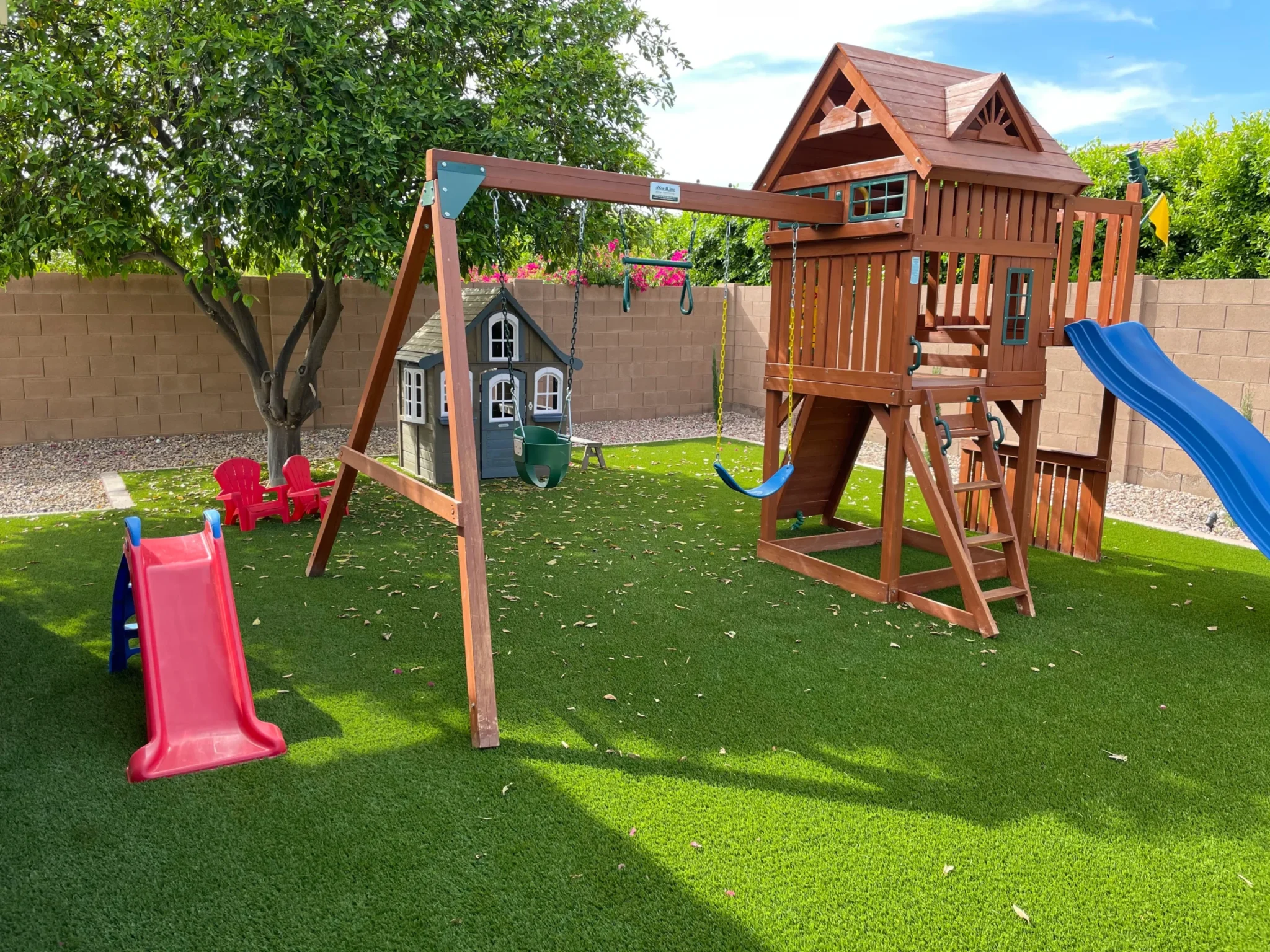 A backyard playground with a wooden playset, slides, swings, a small playhouse, two red kids' chairs, and artificial grass, surrounded by a brick wall and trees.