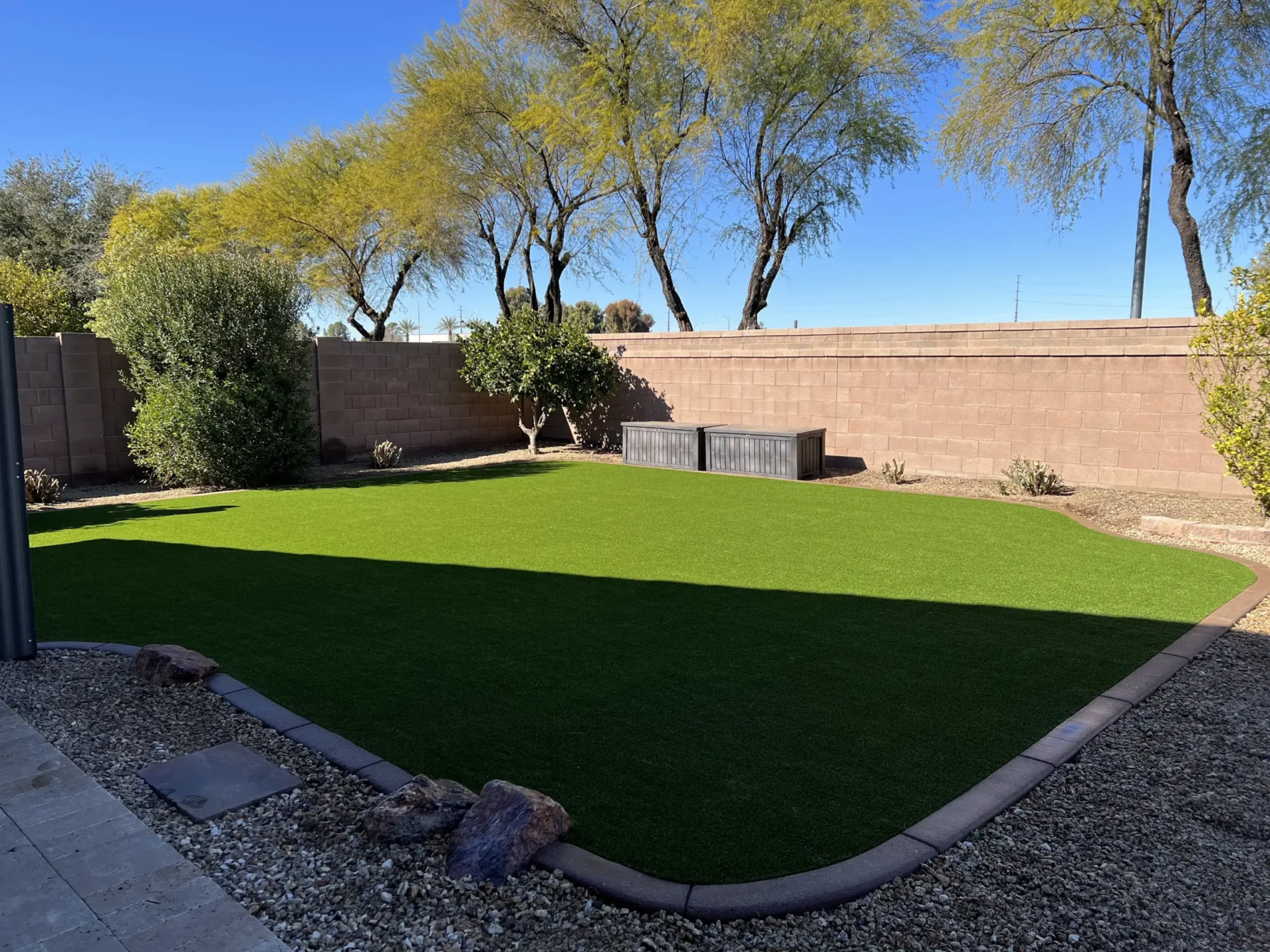 A Texas backyard with artificial grass, bordered by rocks and gravel and enclosed by a tan brick wall. Trees and bushes line the fence, and a storage bench sits against the wall under a clear blue sky.