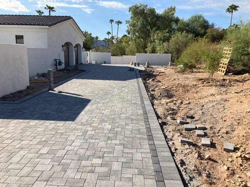 A backyard swimming pool with light-colored tile and expertly designed pavers installed by a trusted Pavers Installer Texas, artificial green grass along the edges, and tan brick walls enclosing the area. A house and trees are visible in the background.