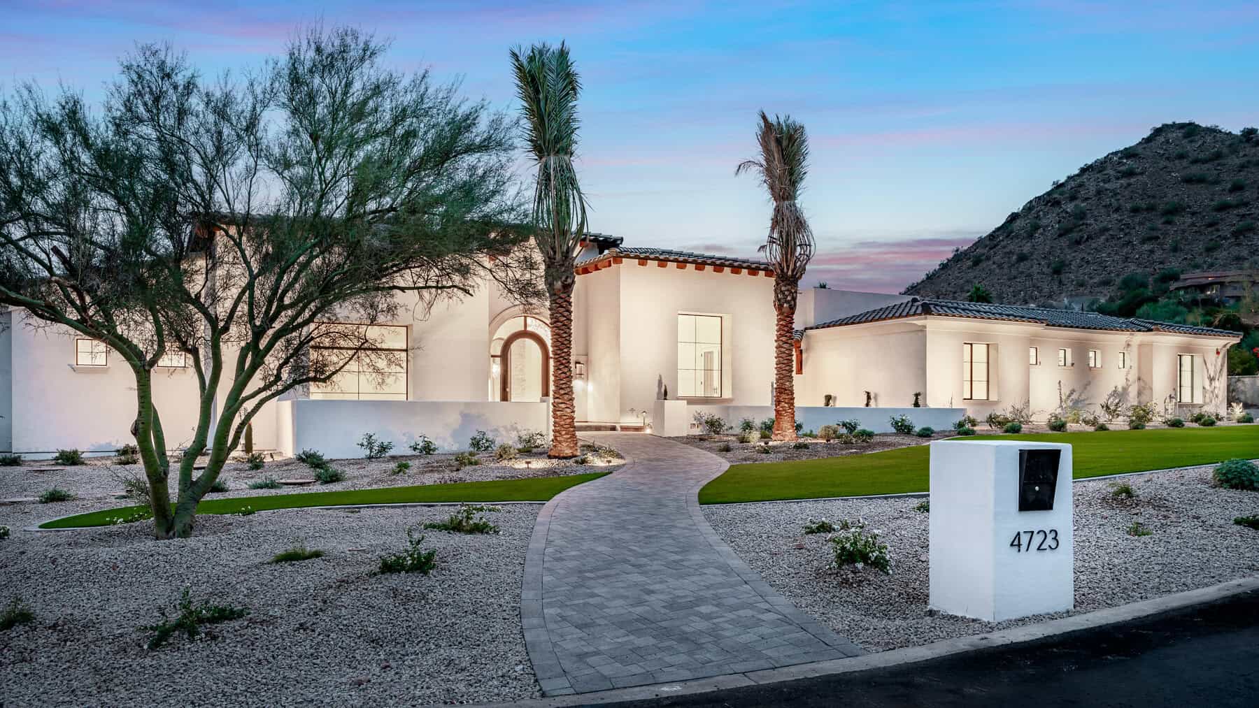 Modern backyard designed by a Pavers Installer Texas, featuring light gray pavers, a rectangular artificial grass lawn, white gravel borders, a low concrete wall, young trees along the fence, and a clear blue sky above.