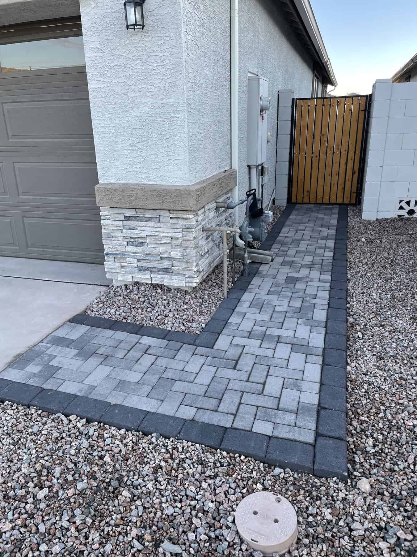 A narrow pathway made of rectangular pavers, expertly crafted by a Pavers Installer Texas, runs alongside a beige house, bordered by gravel on both sides and a bush near a block wall fence.