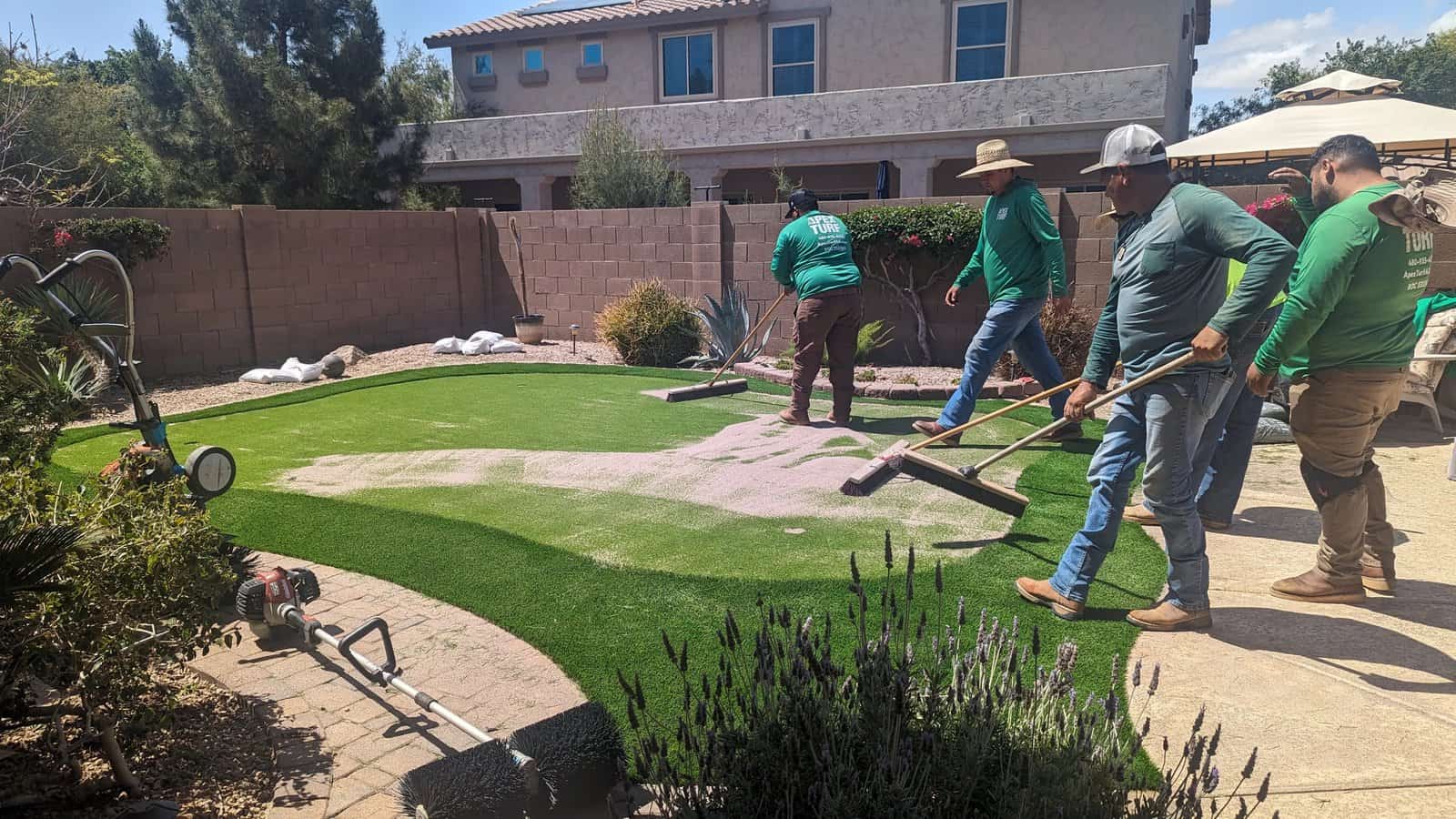 A backyard putting green with four small flag poles, surrounded by artificial grass, desert plants, and a beige brick wall creates the perfect spot to practice your golf short game under a clear blue sky.