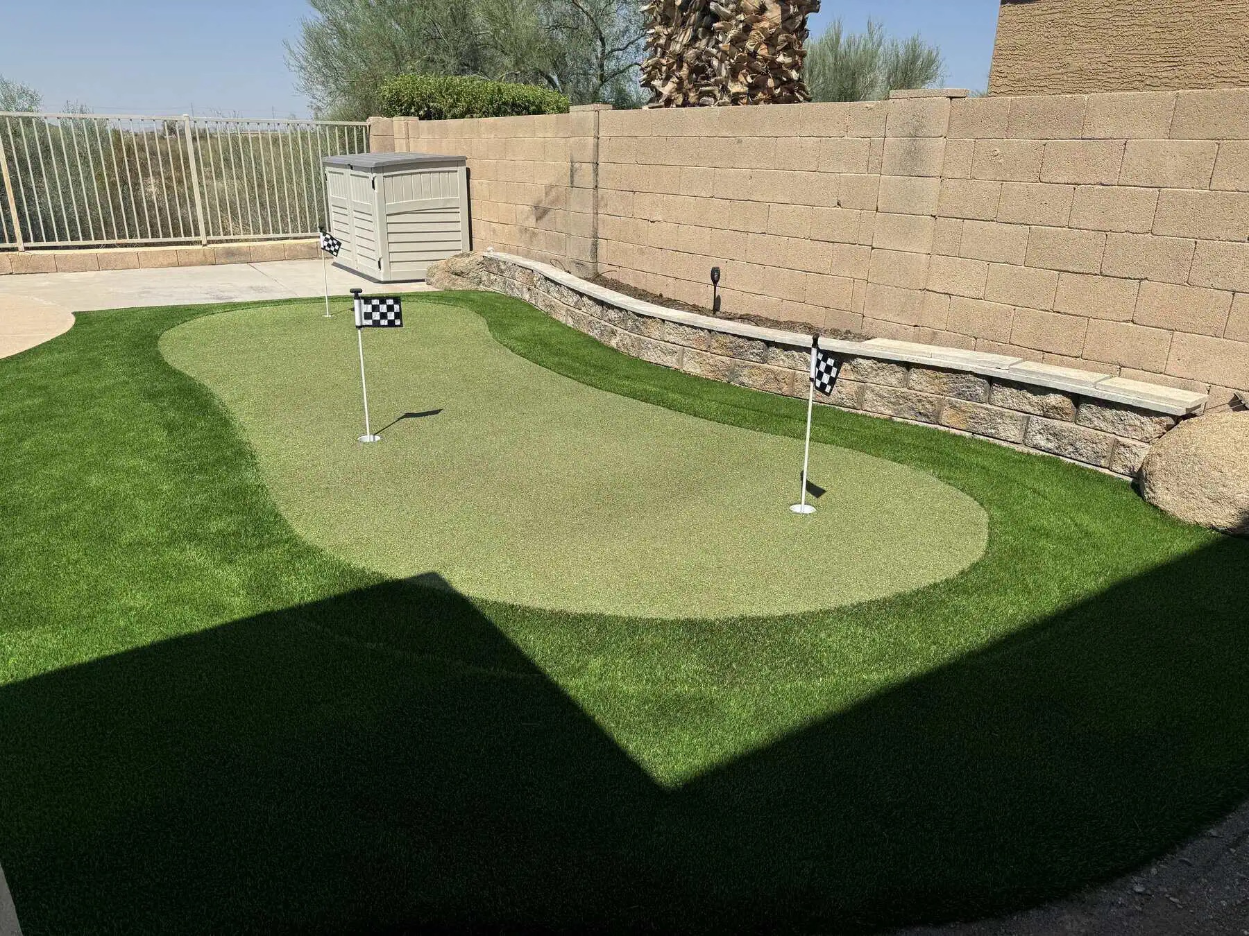 A Texas backyard with artificial grass, bordered by rocks and gravel and enclosed by a tan brick wall. Trees and bushes line the fence, and a storage bench sits against the wall under a clear blue sky.
