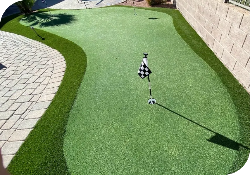 A backyard putting green with artificial turf, golf flags, large rocks, a white sand trap, and lush landscaping, all set against a tall tan wall and adjacent to a nearby patio area.
